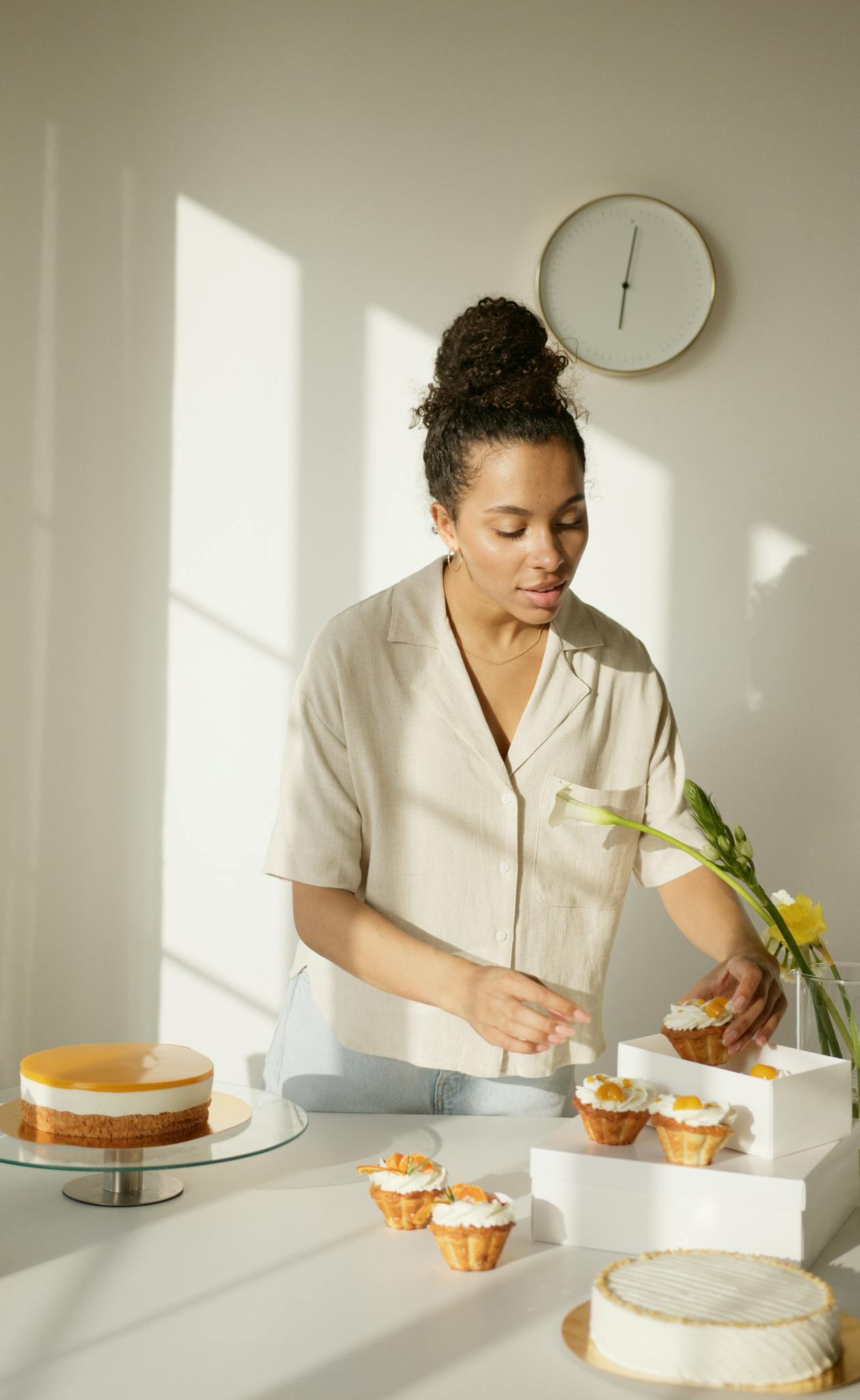 A woman in a sunlit room arranging cupcakes alongside cakes, displaying a fresh homemade vibe.