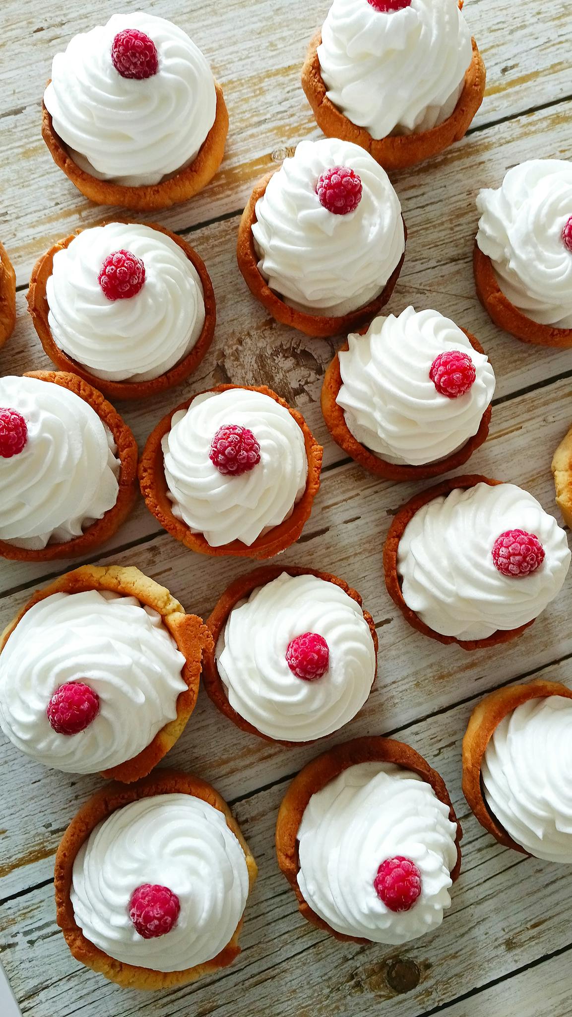 Flat lay of raspberry tartlets topped with cream on a wooden surface.